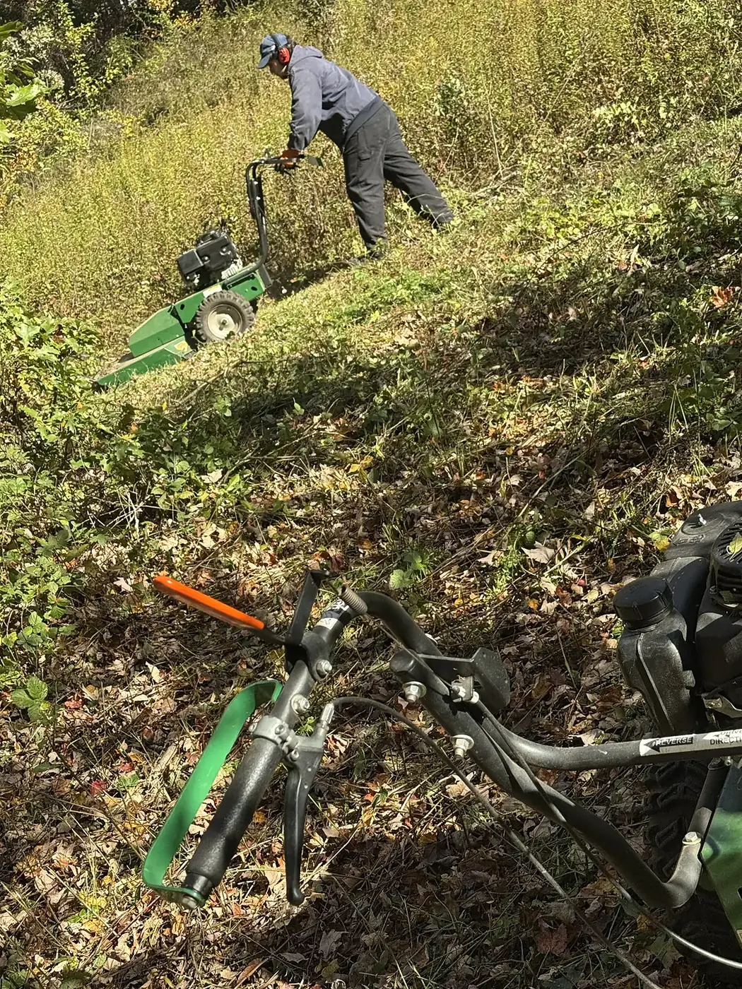 Brush mower in the foreground while a crew member clears a slope
